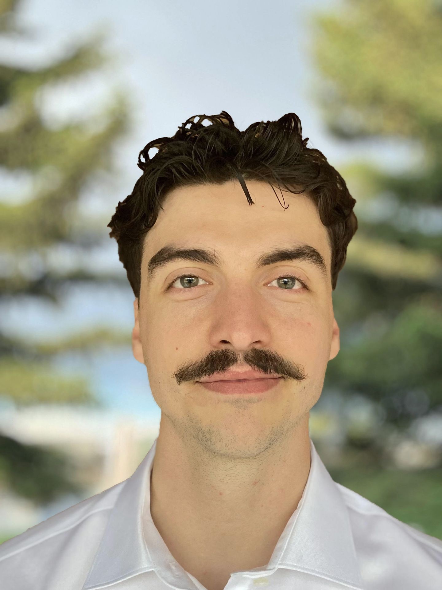 Man with a mustache wearing a white shirt outdoors, with blurred trees in the background.